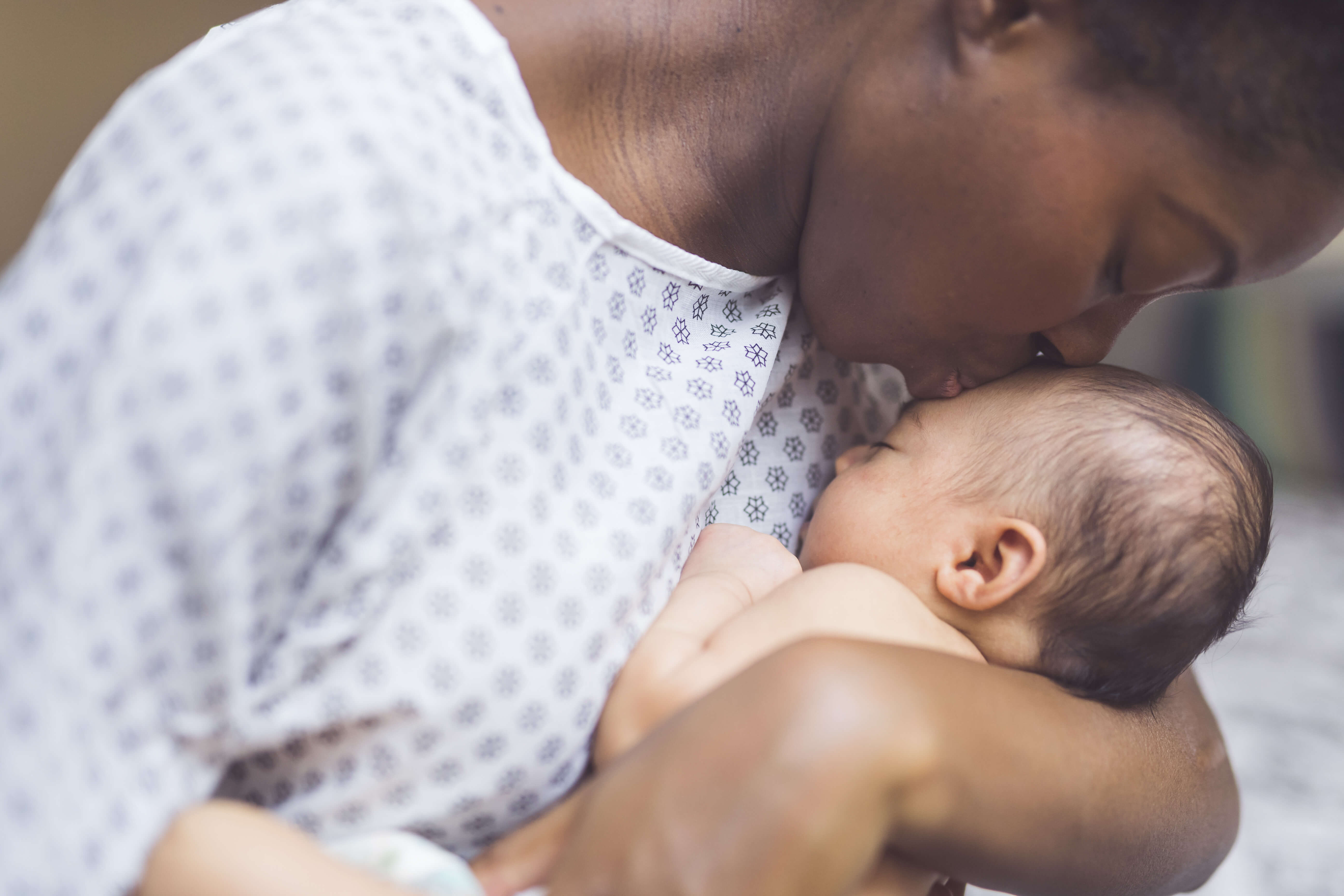 A beautiful young African American mother in a hospital gown gently holds her infant in her arms and smiles down at her. The baby's eyes are closed.