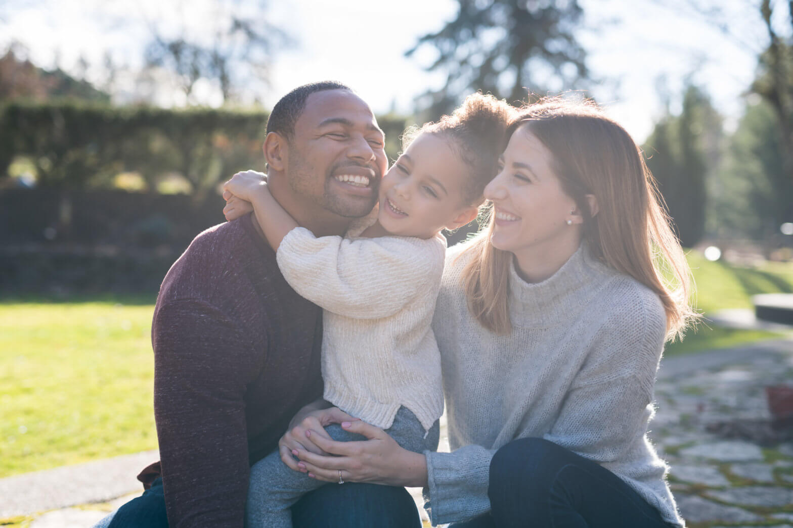 Portrait of a pre-school age girl and her parents sitting outside i the sunshine. Mom is white, dad is black, and they are all happy and affectionate as they laugh and smile at one another. They are sitting on a curb and there is a grassy area in the background.