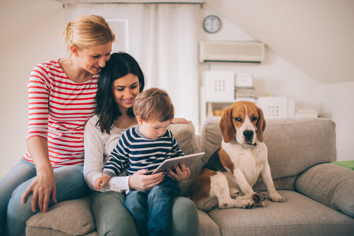 Photo of family, two women, dog and cute little boy, are sitting on the sofa in their living room and using tablet
