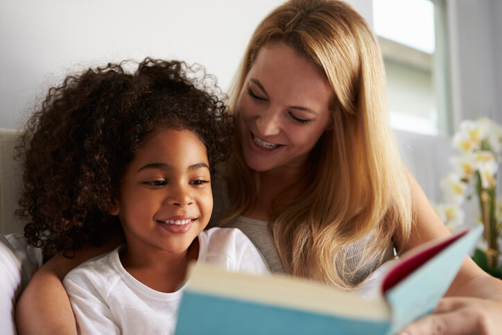 Caucasian mum and smiling black daughter read in bed, close-up