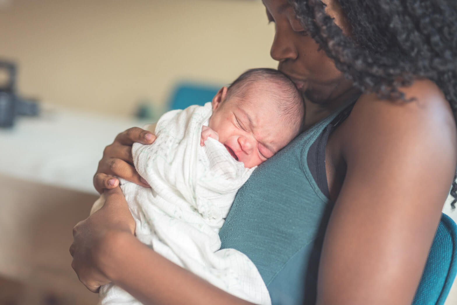 An African American mom gently cradles her newborn daughter against her chest. She is wrapped up and swaddled and is sleeping with a content expression. Mom is tenderly gazing down at her and kissing her head.